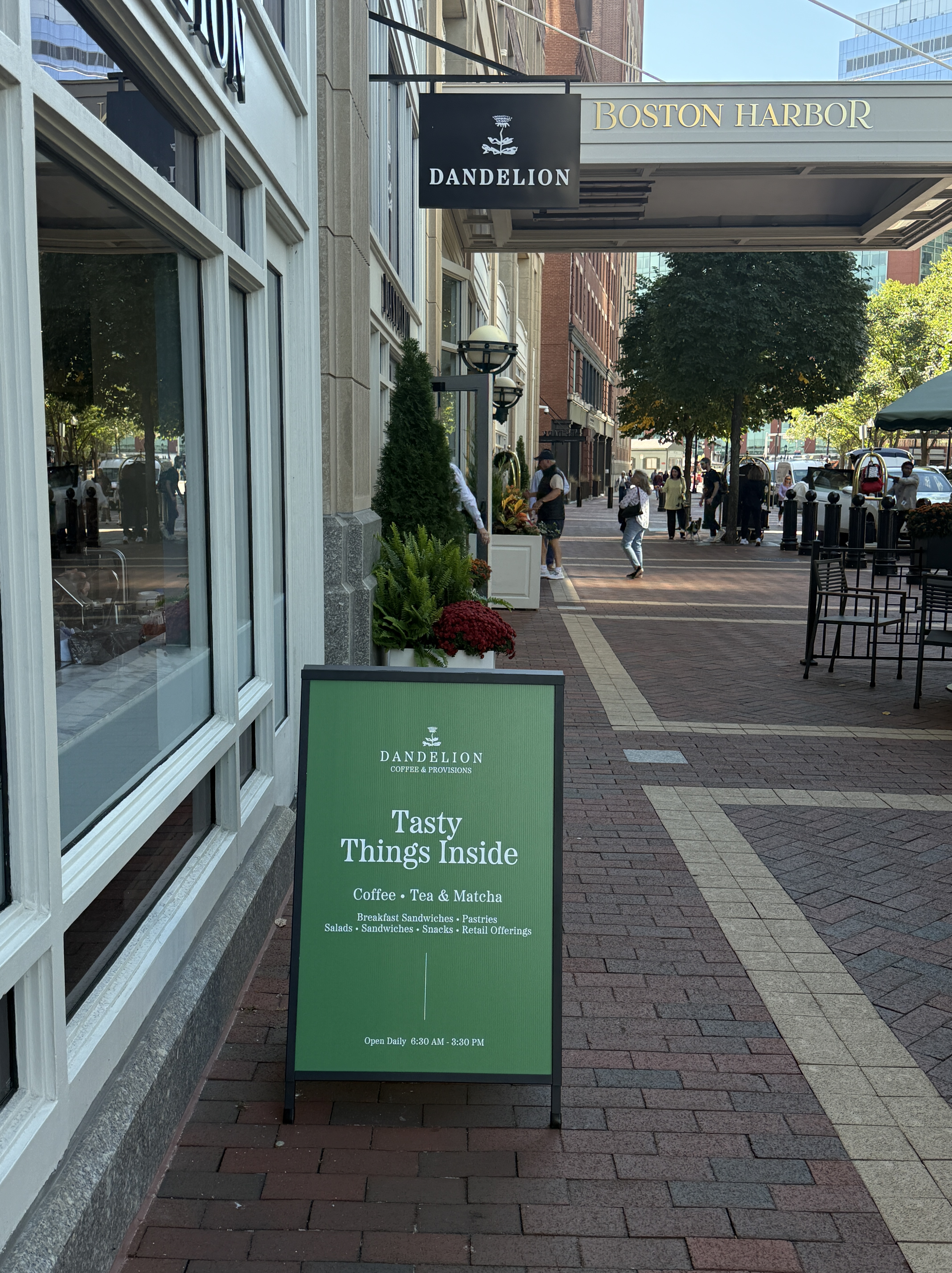 A green A-frame sign outside Dandelion Coffee & Provisions at Boston Harbor Hotel. The sign reads “Tasty Things Inside” with offerings such as coffee, tea, matcha, sandwiches, pastries, and more.