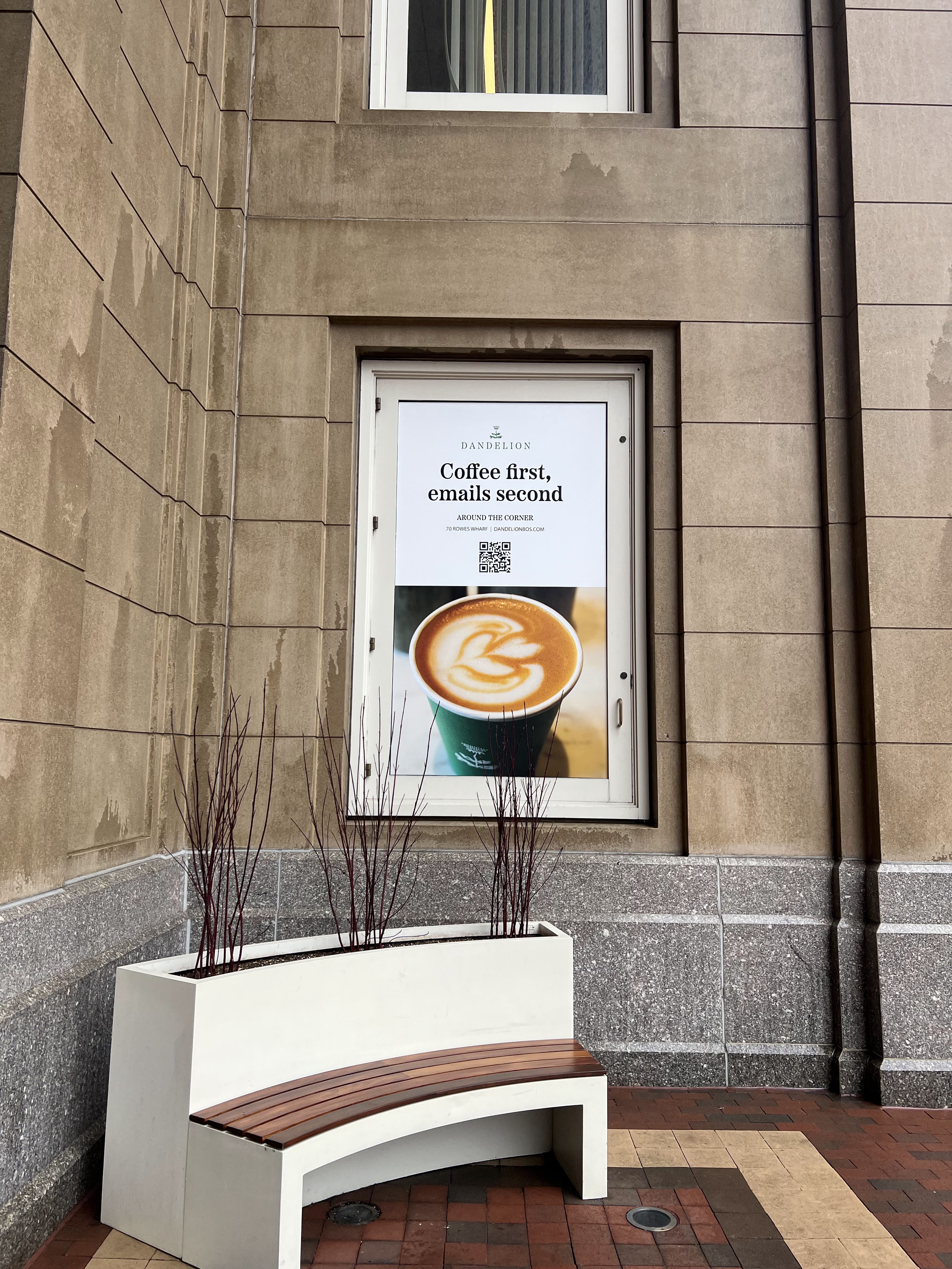 A framed Dandelion Coffee & Provisions poster on the exterior of a stone building, showing a latte and the phrase “Coffee first, emails second” above a QR code. A curved white planter bench sits below.