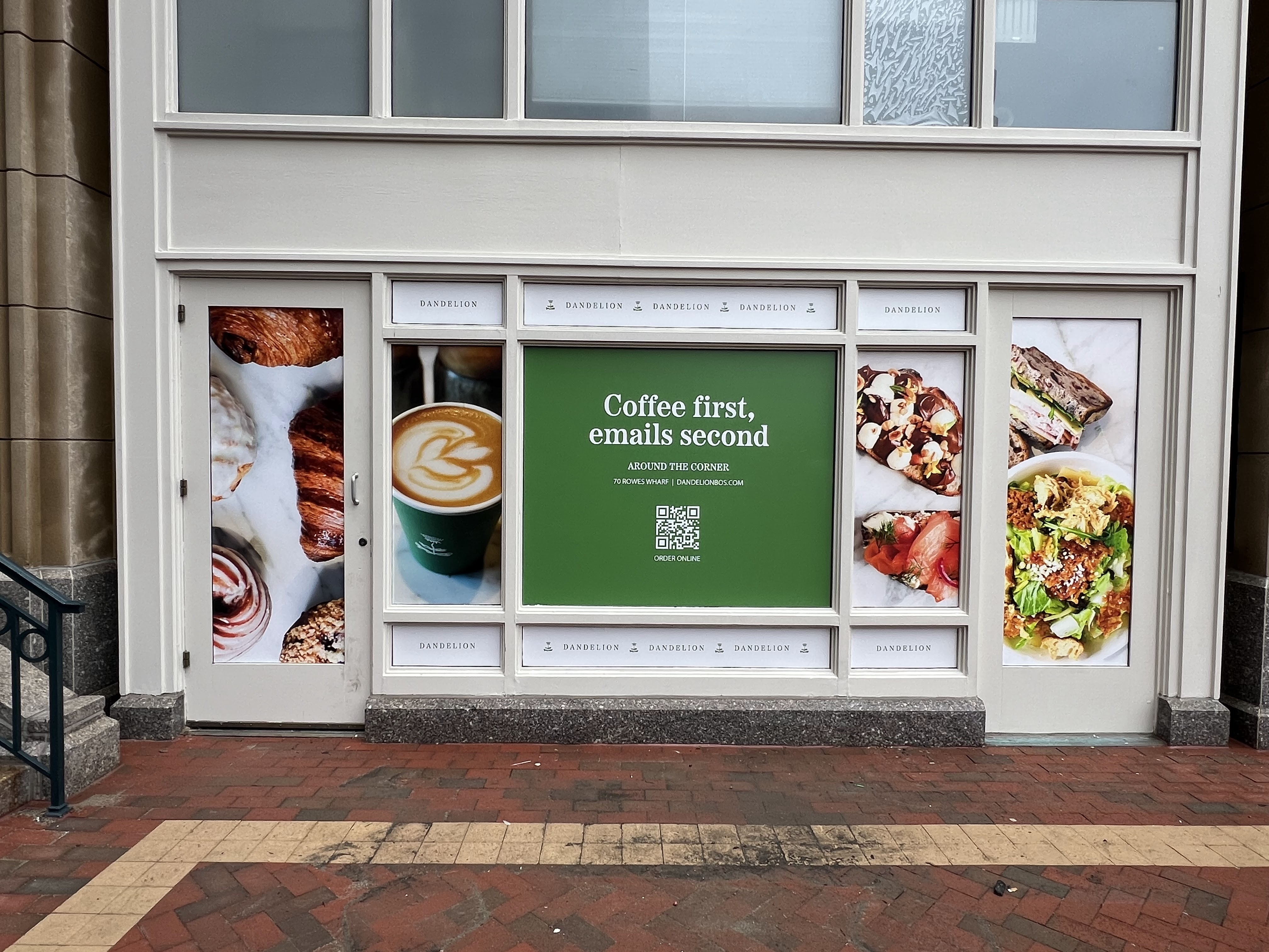 A storefront wrapped with Dandelion Coffee & Provisions advertising, featuring large images of pastries, a latte, salads, and bowls. The center panel displays a green sign reading “Coffee first, emails second” with a QR code for ordering online.
