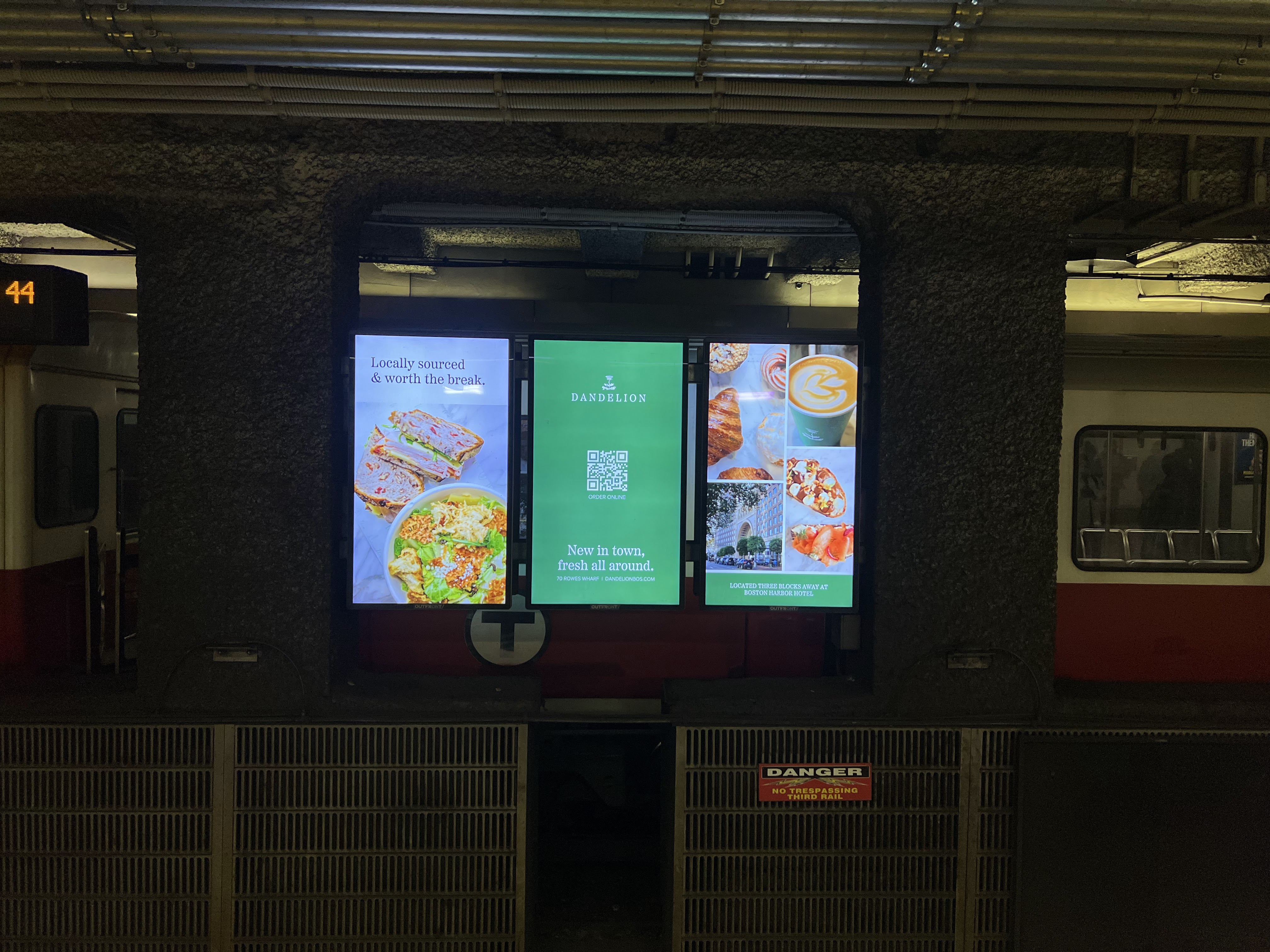 A digital three-panel display inside an MBTA station showing Dandelion advertisements: fresh salad and sandwich imagery on the left, a central green branding panel with QR code, and pastry and latte images on the right.